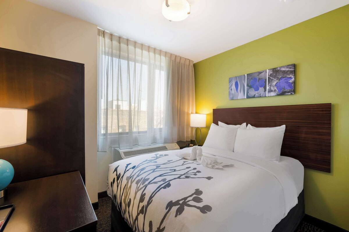 A hotel room with a bed, a desk, a lamp, a window with curtains, and artwork on the wall.