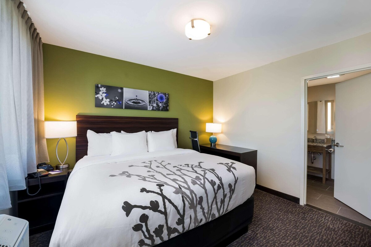 A hotel room with a bed, two bedside lamps, a nightstand, a wall-mounted artwork, and a door leading to a bathroom.