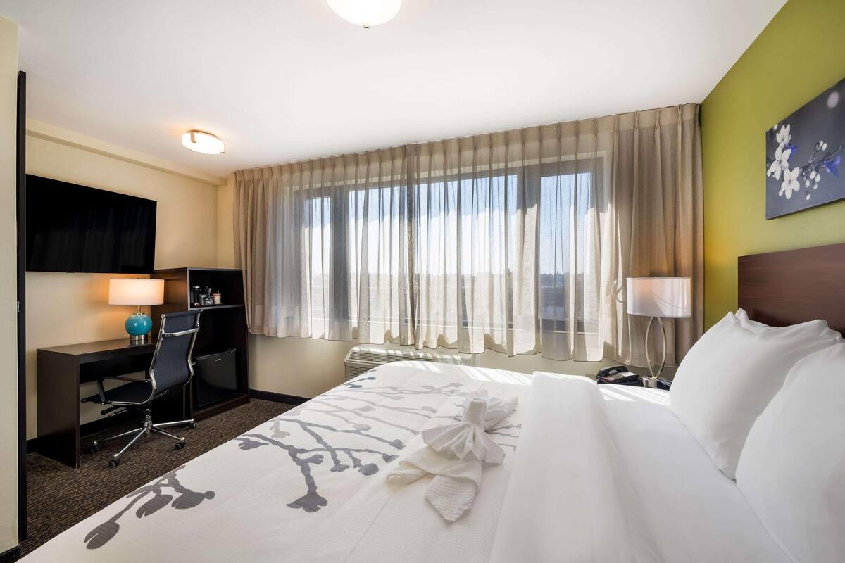 A hotel room with a bed, desk, chair, and a large window with curtains.