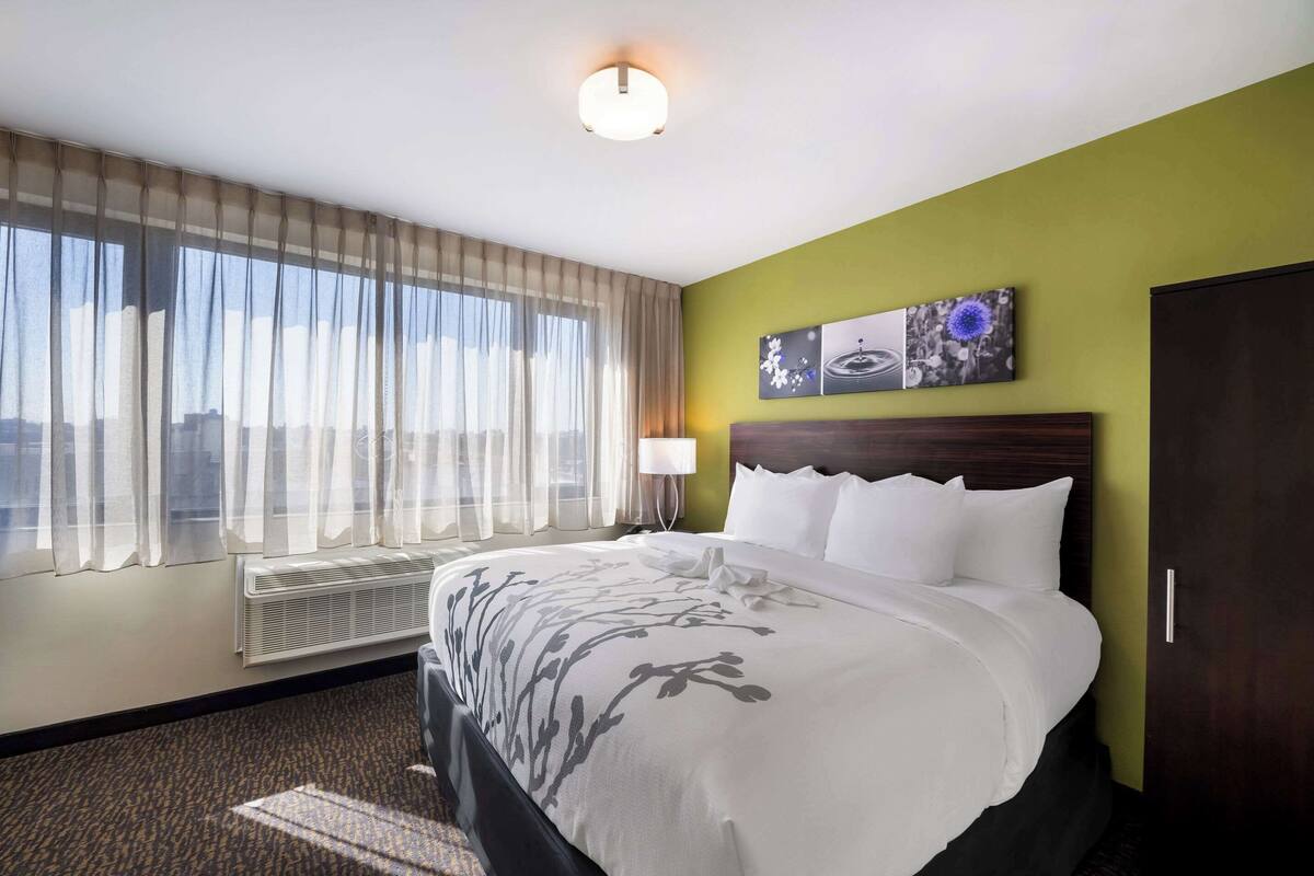 A hotel room with a large bed, a window with sheer curtains, a wall-mounted air conditioner, and a painting above the bed.