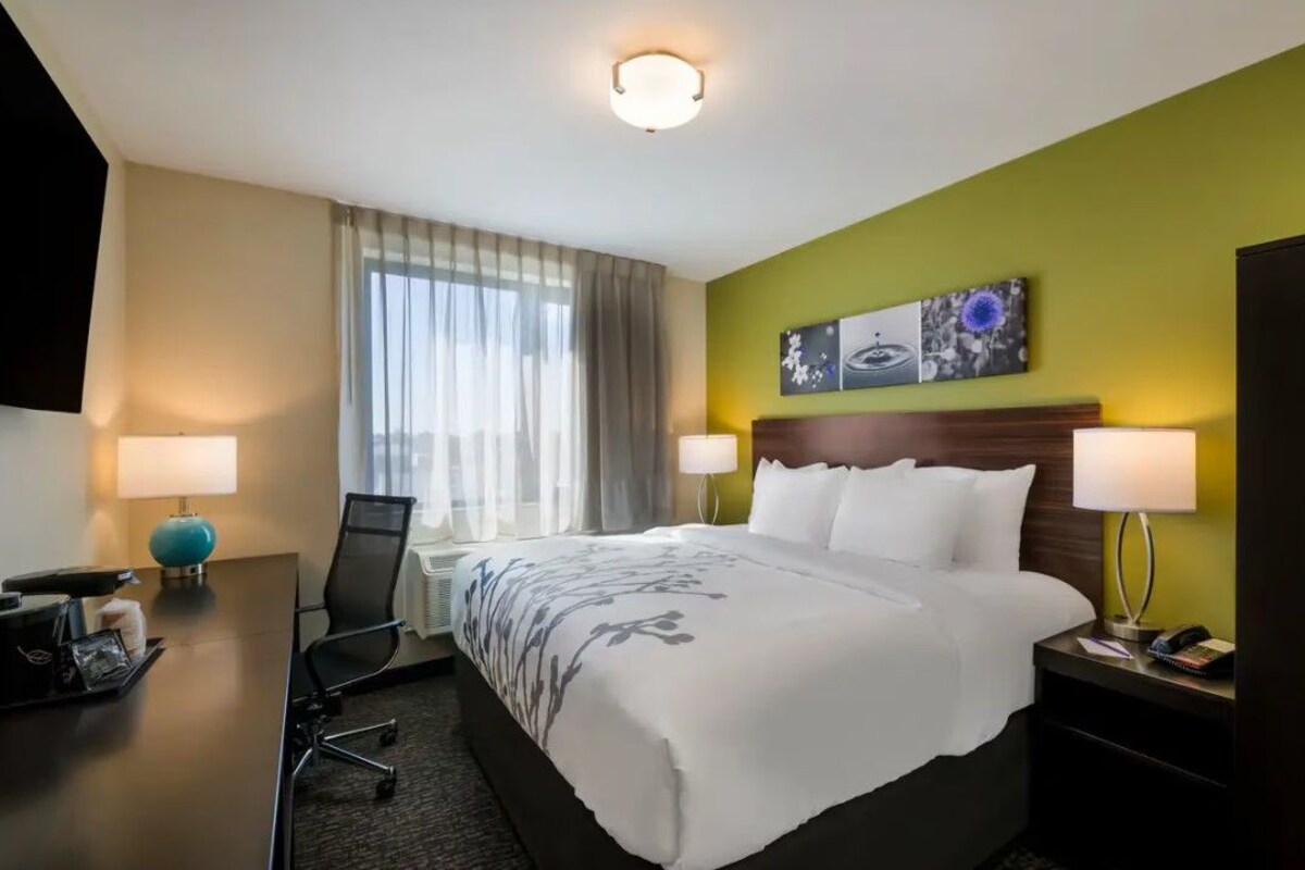 Habitación de hotel con cama, dos lámparas de noche, un escritorio con lámpara, una silla y un ventanal con cortinas.