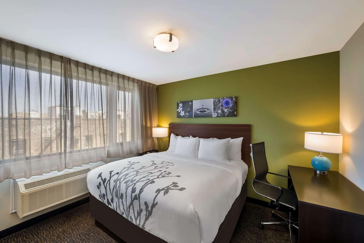 A hotel room with a bed, a desk, a chair, a window with sheer curtains, a wall-mounted lamp, and a ceiling light.
