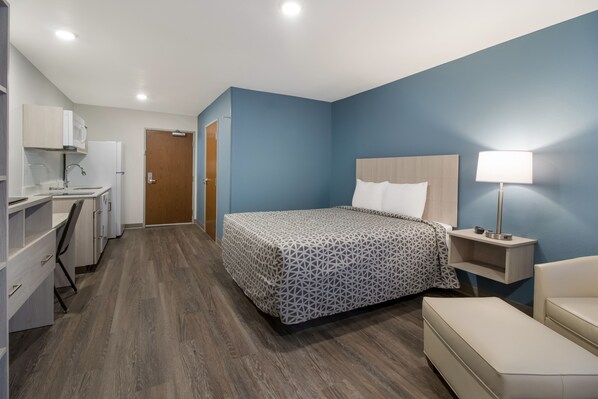 A hotel room with a bed, bedside table, lamp, and a kitchenette area.