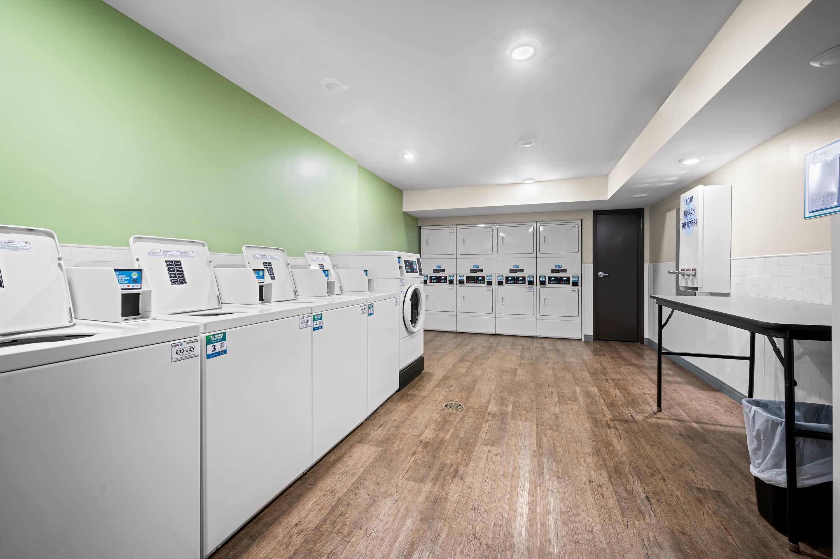 A laundry facility with multiple washing machines and dryers, a wooden floor, and a green wall.
