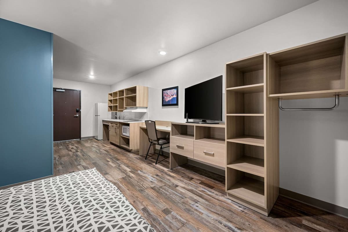 A modern, well-lit room with a wooden floor, a desk with a chair, a TV mounted on the wall, and built-in shelving.