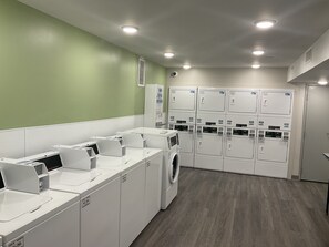 A laundry facility with multiple washing machines and dryers, a green wall, and a wooden floor. - Woodspring Suites Dearborn - Detroit (Dearborn)