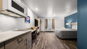 A compact hotel room with a kitchenette, a bed with a patterned bedspread, a desk with a chair, and a window with curtains. - Woodspring Suites Dearborn - Detroit (Dearborn)