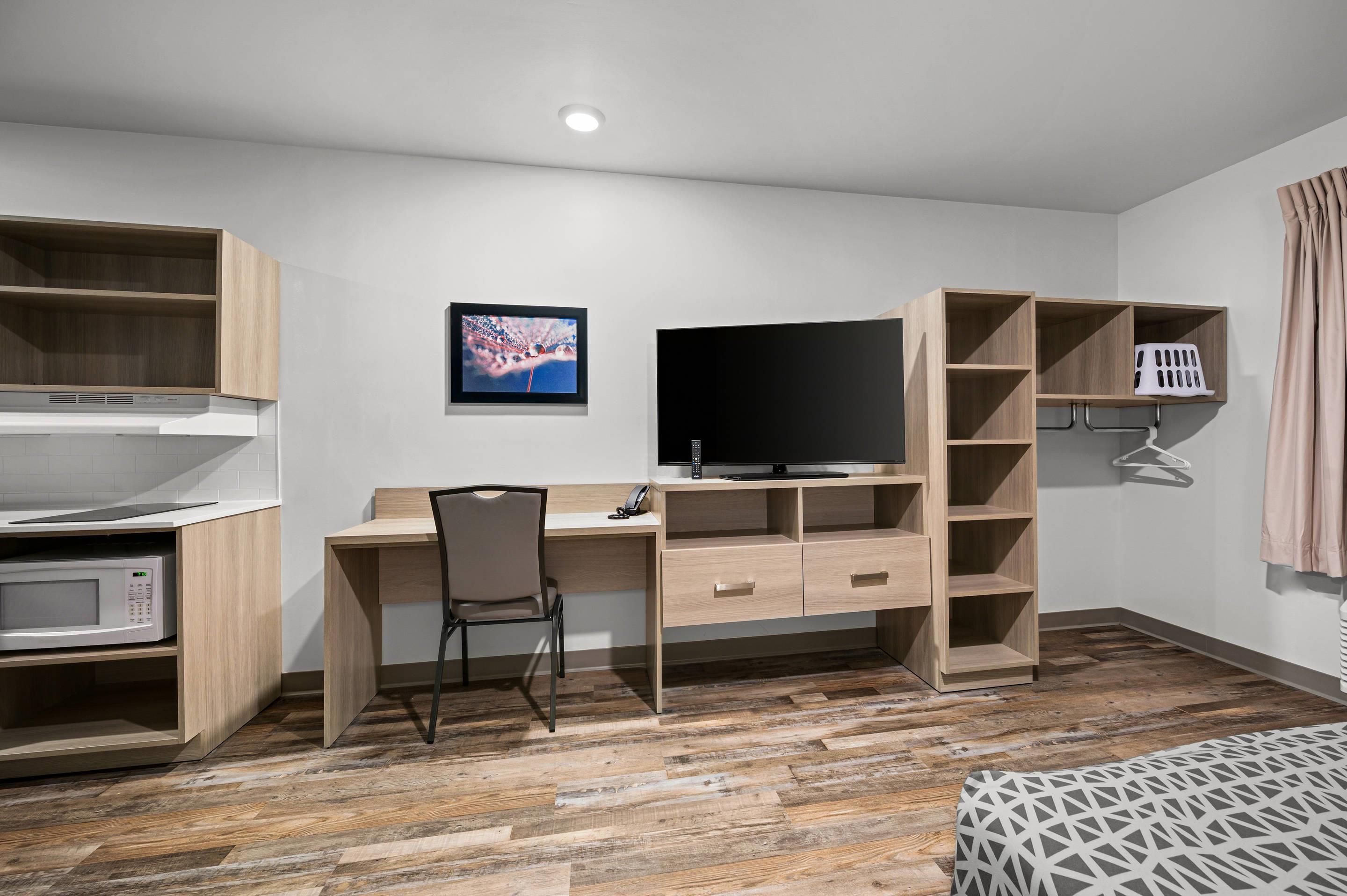 A compact hotel room with a kitchenette, a desk with a chair, a flat-screen TV, and a wardrobe with hanging space.