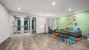 A modern living room with a gray sofa, a coffee table, and a colorful rug. There are glass doors leading to another room, a floor lamp, and framed pictures on the wall. - Woodspring Suites Dearborn - Detroit (Dearborn)