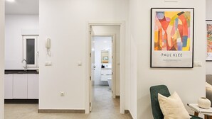 Un interior moderno con un pasillo que conduce a una cocina y una sala de estar. Hay una obra de arte enmarcada en la pared y un sillón verde con cojín.