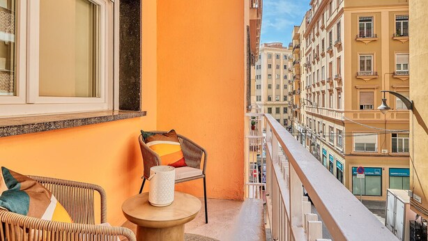 Un balcón con mobiliario de mimbre, una mesa pequeña y una ventana con vistas a la ciudad.