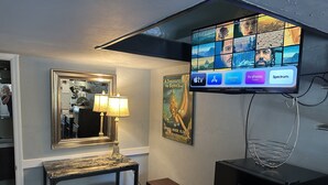 A compact living space with a television mounted on the wall displaying various channels, a small table with two stools, a decorative lamp, and a framed poster.