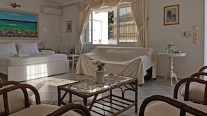 Ein gut beleuchteter, geräumiger Raum mit einem Bett, einer gepolsterten Couch, einem Esstisch mit Stühlen und Blick ins Freie durch große Fenster.