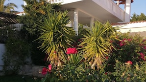 Ein weißes Gebäude mit Balkon, umgeben von üppigem Grün und blühenden Pflanzen.