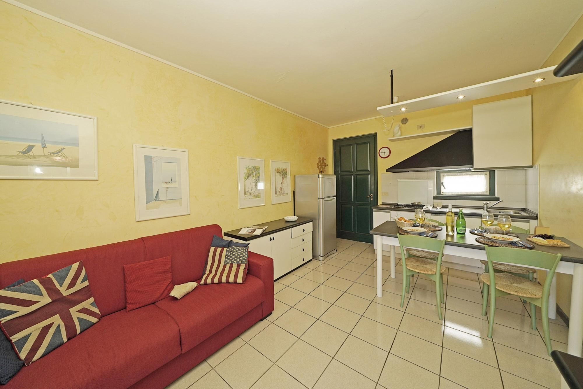 A compact living space with a red sofa, a dining table with chairs, a kitchen area, and a small refrigerator.