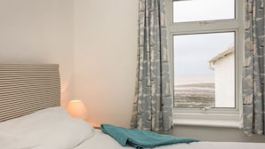A bedroom with a bed, a lamp, and a window with curtains. - Oyster Catchers (Watchet)