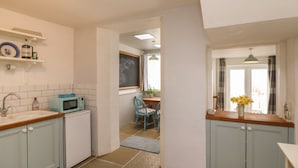 A kitchen with light blue cabinets, a microwave, and a sink. There is a window with curtains and a door leading to another room. - Oyster Catchers (Watchet)