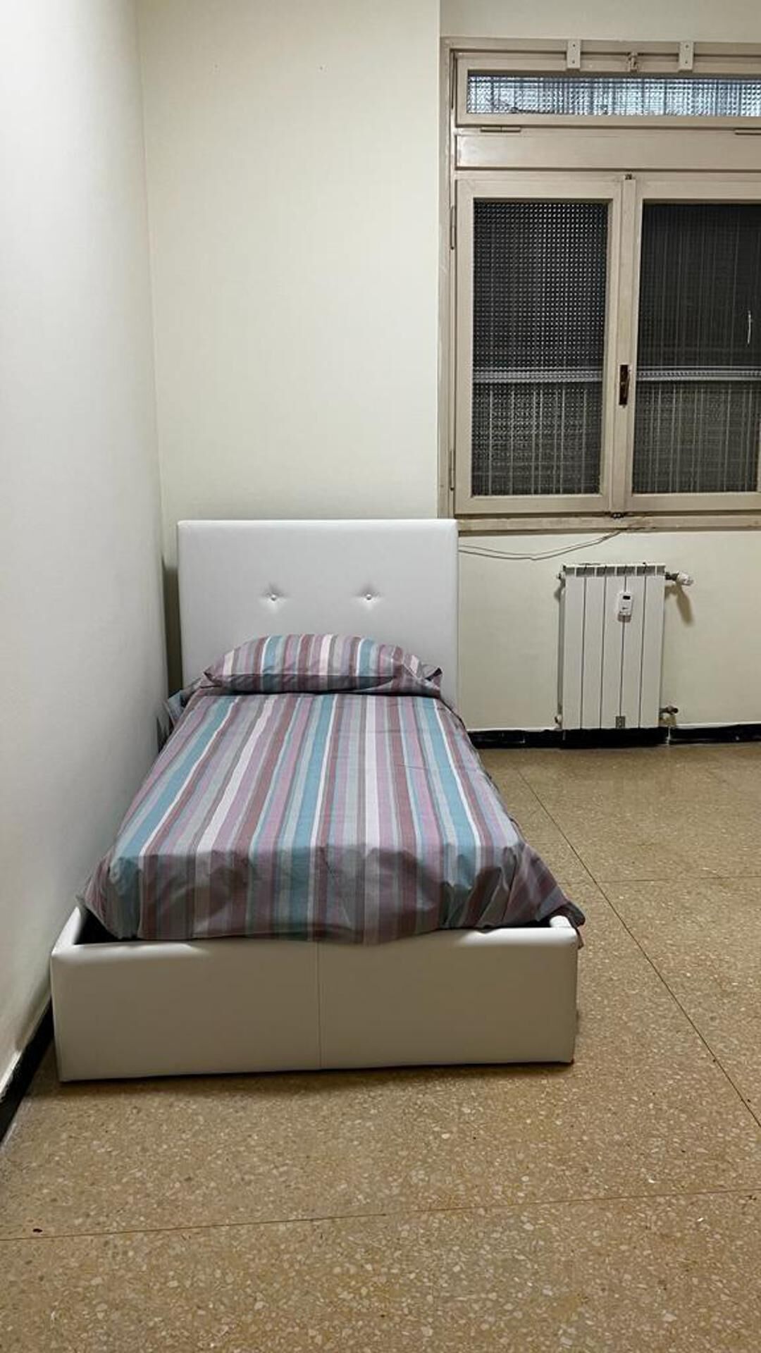 Ein kleines Zimmer mit einem Einzelbett, einem weißen Kopfteil und einer gestreiften Bettdecke.