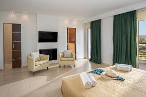 Habitación de hotel moderna con cama, dos sillones, televisor de pantalla plana, chimenea y un ventanal grande con cortinas.