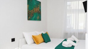 Een hotelkamer met een wit bed, een groene plaid en een witte nachtkastje met een ringlamp.