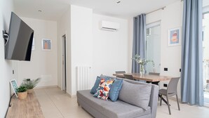 Een moderne woonkamer met een grijze bank, een houten bijzettafeltje, een flatscreen televisie en een eethoek met een houten tafel en stoelen.