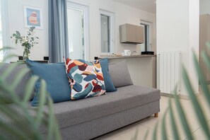 A modern living room with a grey sofa, colorful cushions, a vase with flowers, and a kitchen in the background.