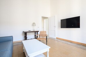 Une pièce avec une table blanche, une chaise en osier, un canapé bleu, une lampe et un téléviseur fixé au mur.