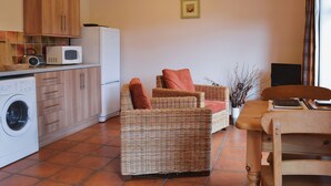 Une cuisine équipée d’un lave-linge, d’un four à micro-ondes et d’un réfrigérateur. On y trouve une chaise en osier avec des coussins, ainsi qu’une table sur laquelle sont posés un livre et une plante.