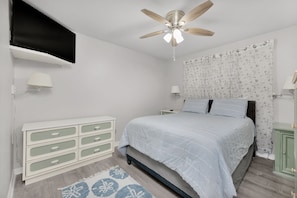 Un dormitorio con una cama, ventilador de techo, cómoda y televisor montado en la pared.