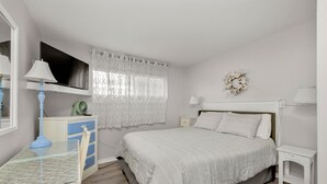 Un dormitorio con cama, mesitas de noche, un escritorio con lámpara, un espejo y una ventana con cortina.