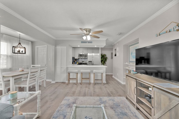 Una cocina moderna con armarios blancos, una isla central con taburetes, un ventilador de techo y un televisor de pantalla plana grande.