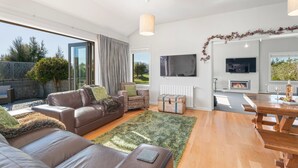 Ein modernes Wohnzimmer mit einer Couch, einem Couchtisch und einem Kamin. Ein Fernseher ist an der Wand befestigt und ein Fenster bietet Blick auf einen Garten.