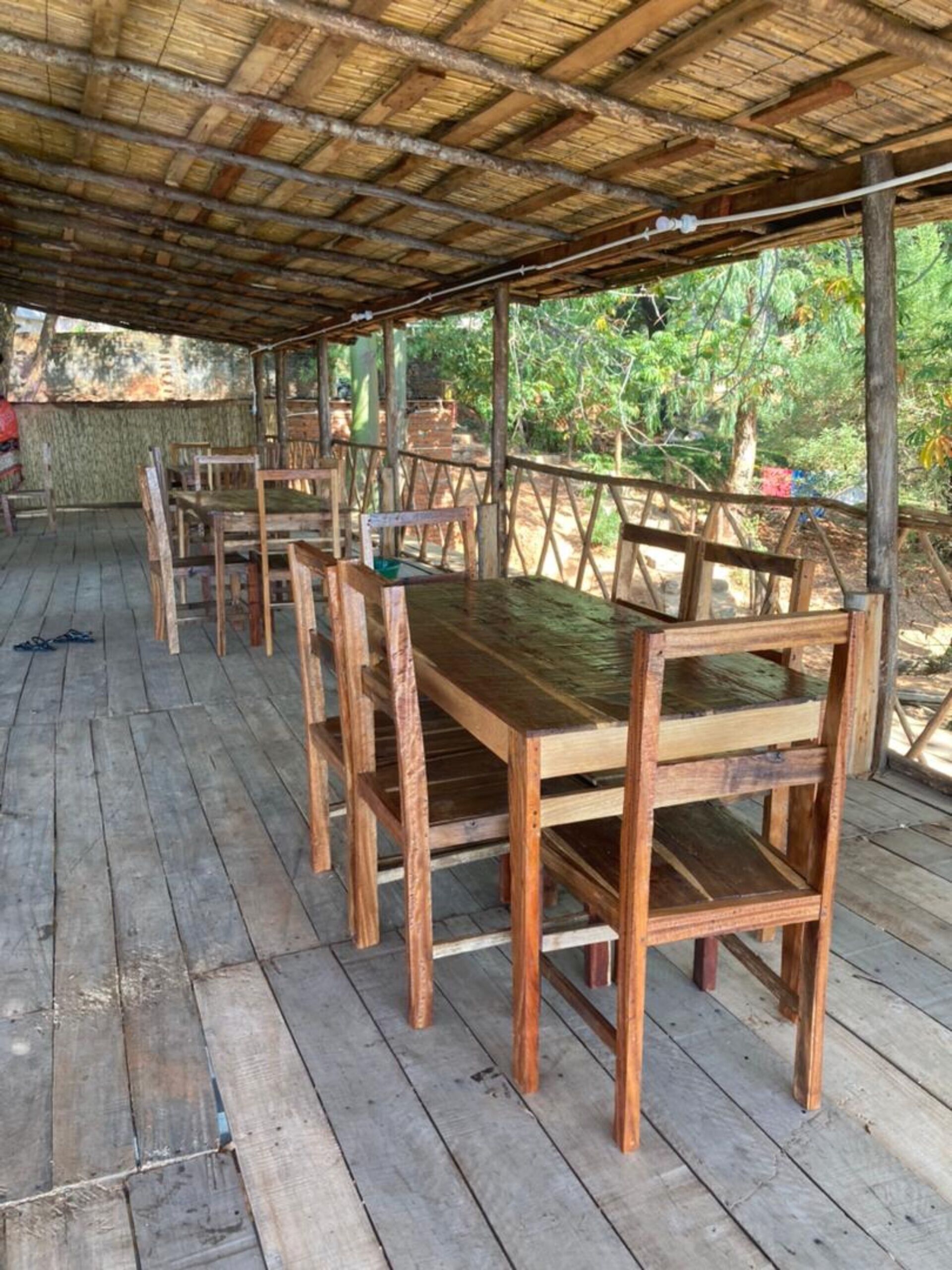 Un espacio de estar al aire libre cubierto, con mesas y sillas de madera, rodeado por una terraza de madera y un techo de paja.