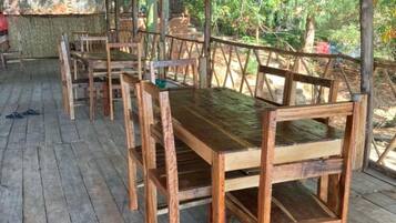 Un espacio de estar al aire libre cubierto, con mesas y sillas de madera, rodeado por una terraza de madera y un techo de paja.