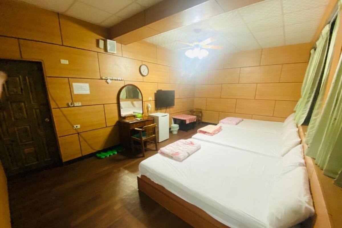 Habitación de hotel con dos camas, piso de madera, ventilador de techo, escritorio con silla, espejo y televisor.