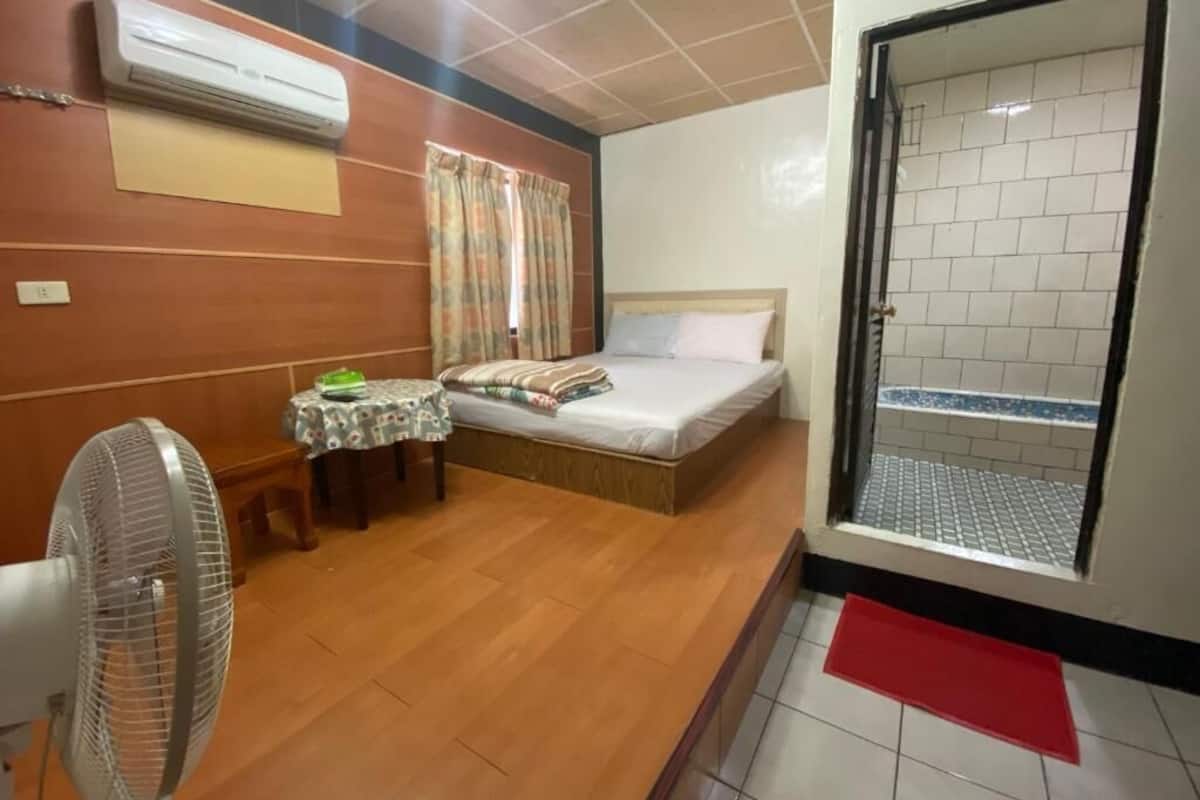 Habitación de hotel con cama, una mesita, ventilador, aire acondicionado y vista a un patio con baldosas.