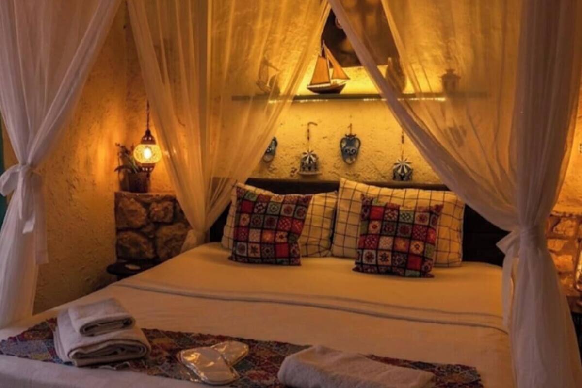 A bed with a canopy, pillows, a nightstand with a lamp, and a wall-mounted shelf.