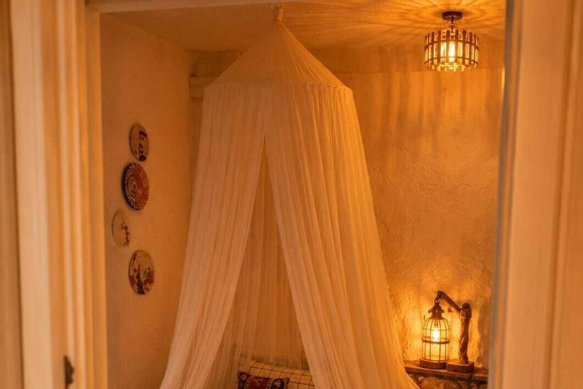 A bedroom with a canopy bed, a stone floor, a nightstand with a lamp, and wall decorations.