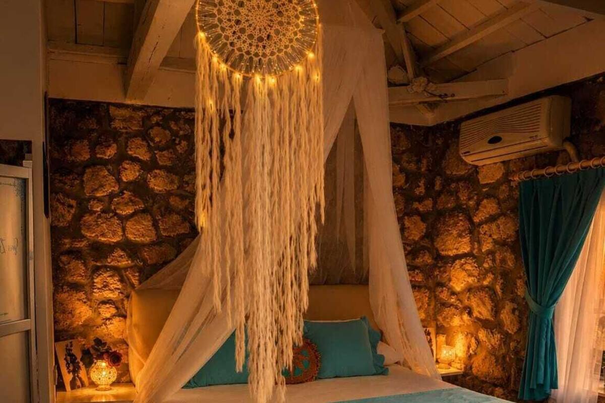 A bedroom with a stone wall, a canopy bed, a bedside table with a lamp, and a window with curtains.