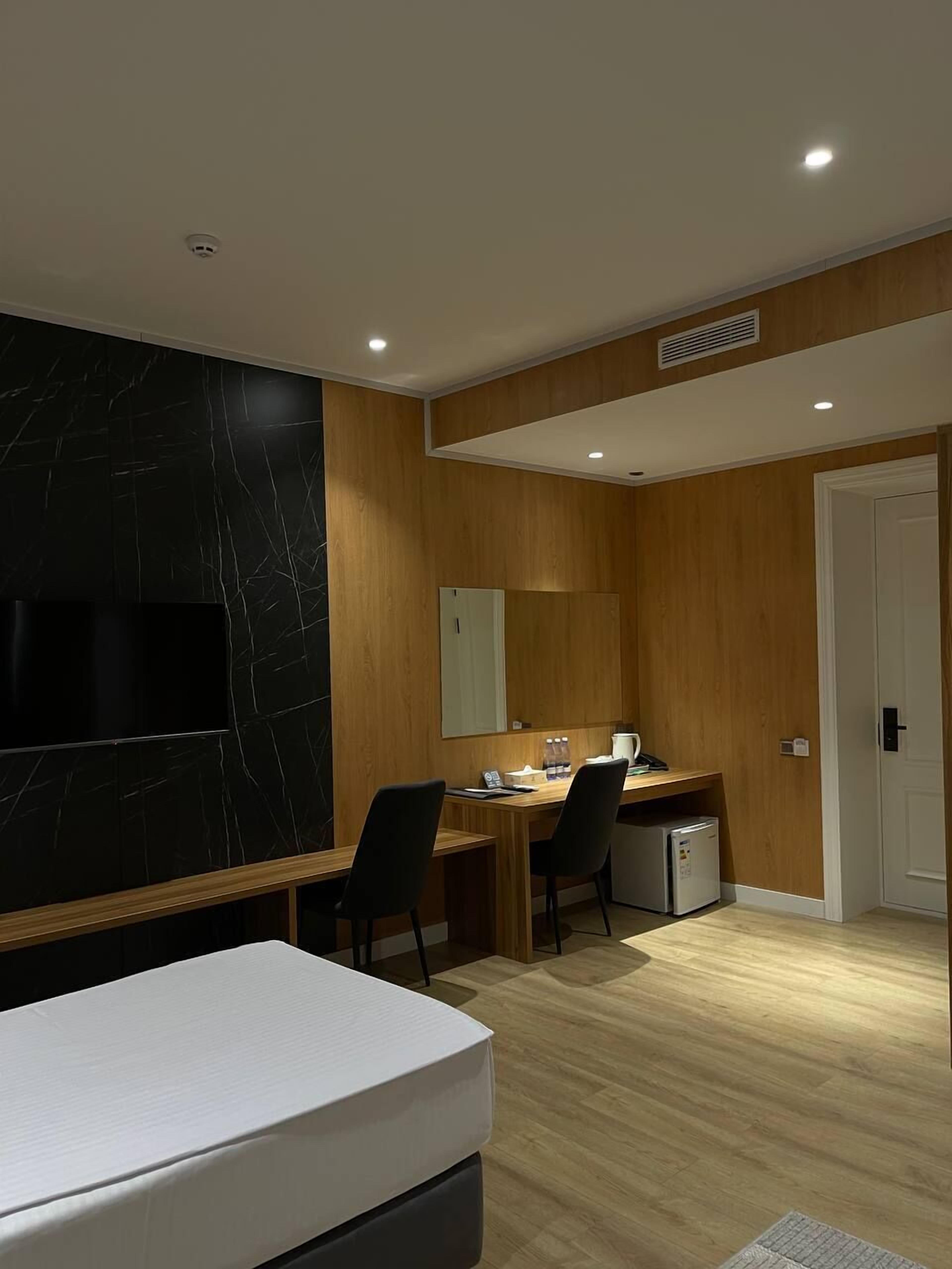 A modern hotel room with a wooden floor, a black marble accent wall, a flat-screen TV, a desk with two chairs, and a small refrigerator.