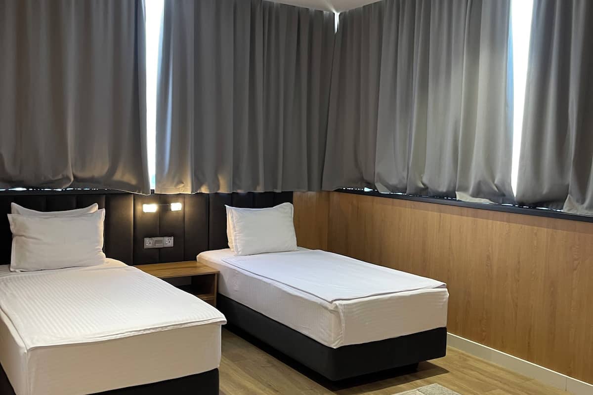 A modern hotel room with two beds, a wooden headboard, a nightstand, and a large window with grey curtains.