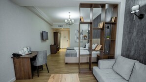 A modern living room with a sofa, a coffee table, a desk, and a bookshelf. There is a bed in the background with a view of the hallway. - WELLFORT HOTEL (Samarkand)