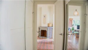 Une porte ouverte donne sur une pièce dont le sol est recouvert de carreaux, qui possède une cheminée décorée et un luminaire suspendu.