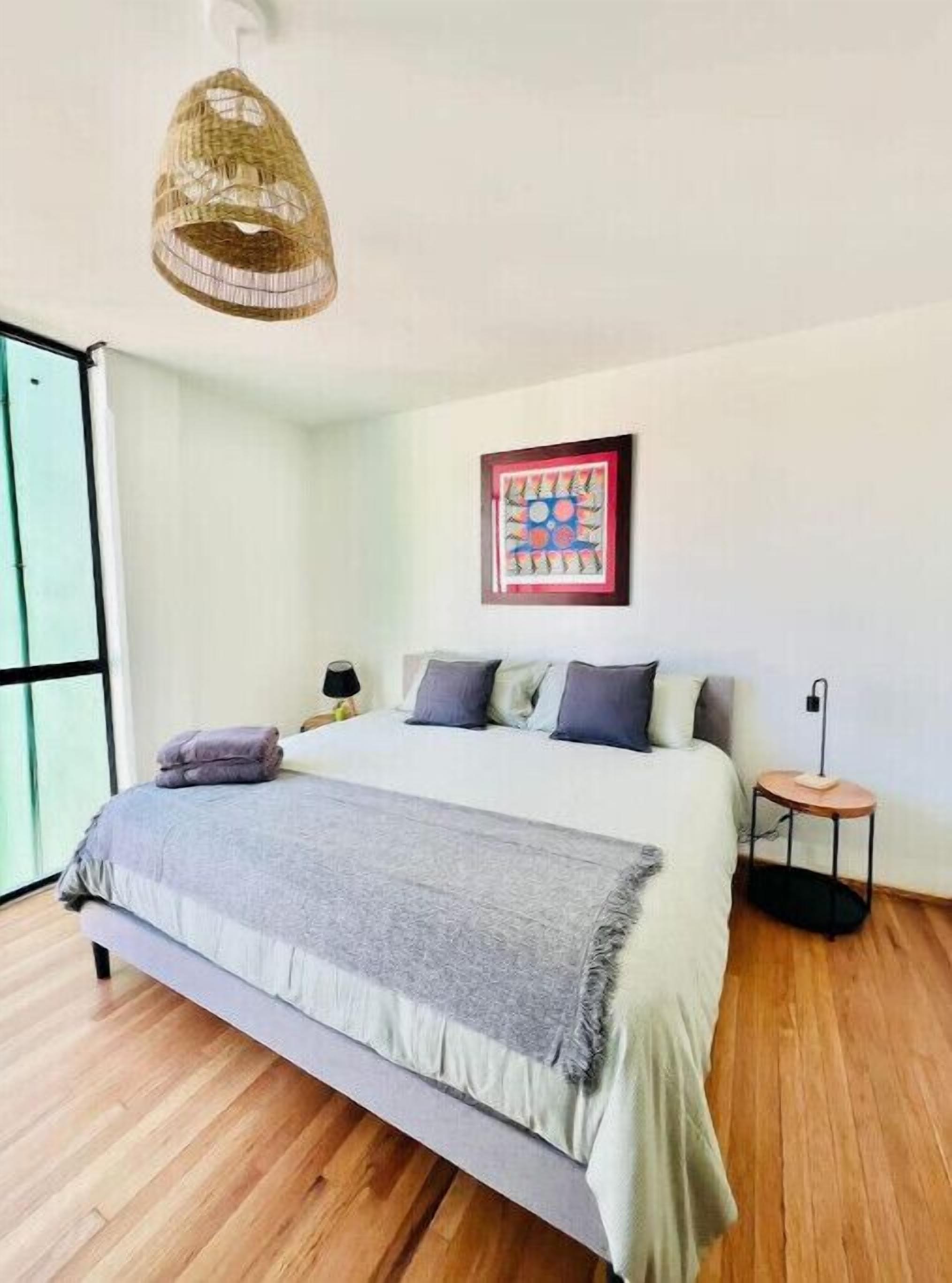 A modern bedroom with a large bed, a hanging light fixture, a framed artwork on the wall, and a small bedside table.