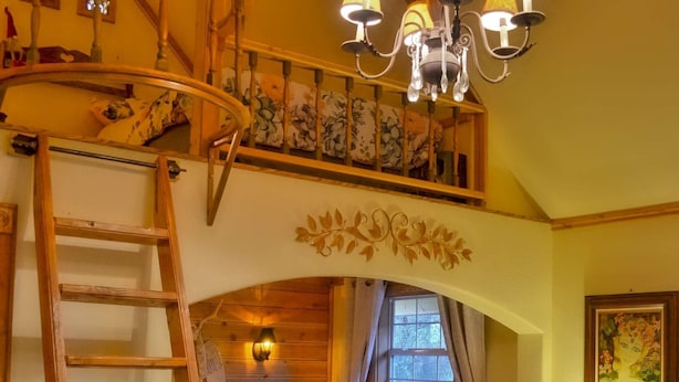 A cozy room with a loft bed, a wooden ladder, a chandelier, and a small table with a lamp. - Schneewittchen Cottage (#3) (Fredericksburg)