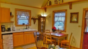 A cozy kitchen with wooden cabinets, a dining table with chairs, a small refrigerator, and a window with lace curtains. - Aschenputtel Cottage (#4) (Fredericksburg)