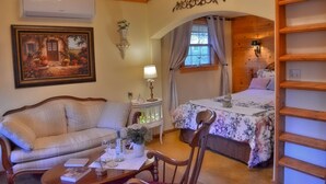 A cozy room with a bed, a sofa, a table with a vase and wine glasses, and a painting on the wall. - Aschenputtel Cottage (#4) (Fredericksburg)