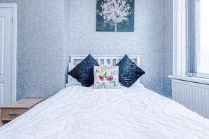Quarto com cama branca, almofadas azul escuras e uma almofada floral. Há uma mesinha de cabeceira branca e uma janela com persianas.