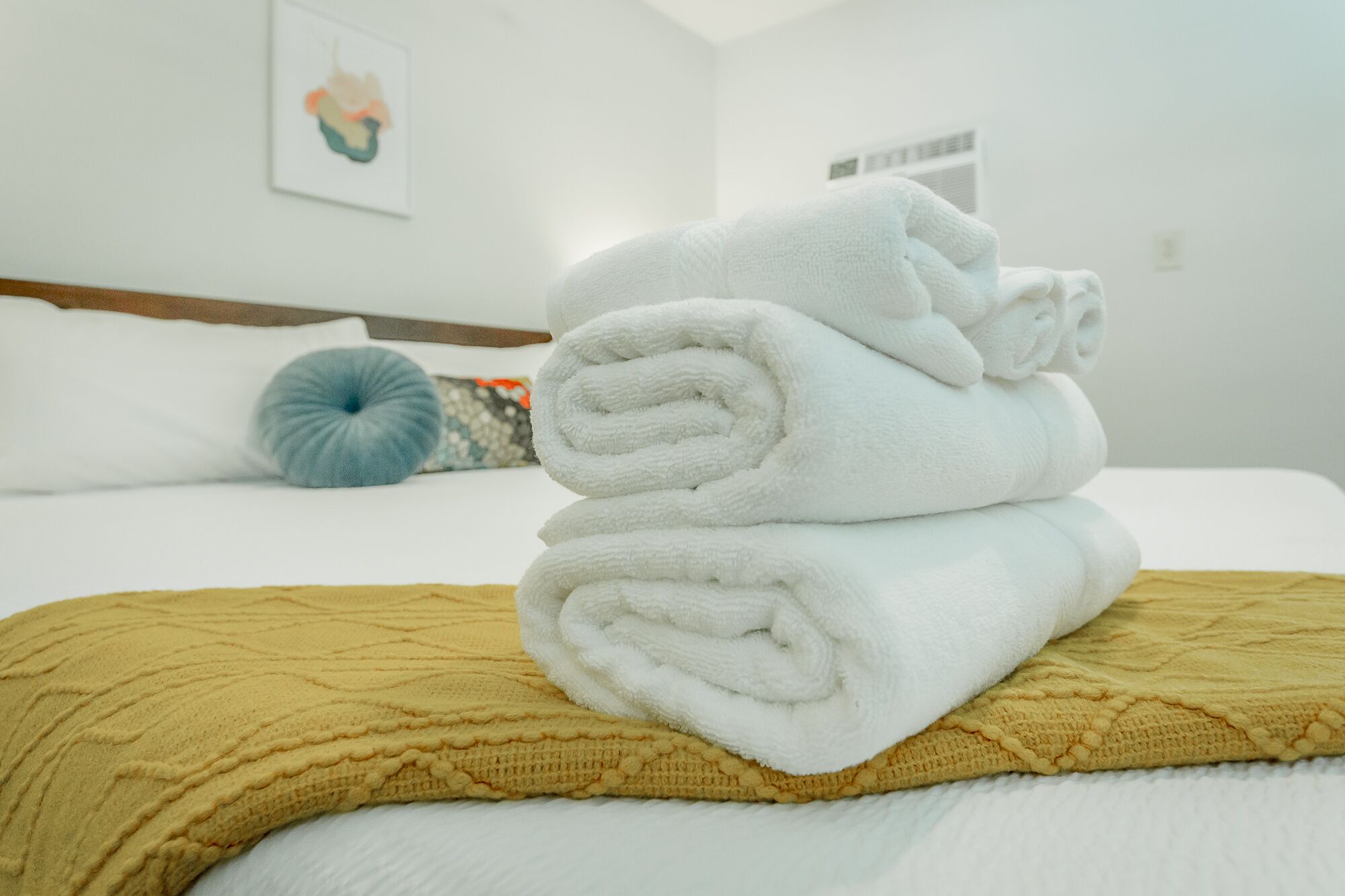 A neatly made bed with white towels, a yellow knitted blanket, and a blue pillow in a hotel room.
