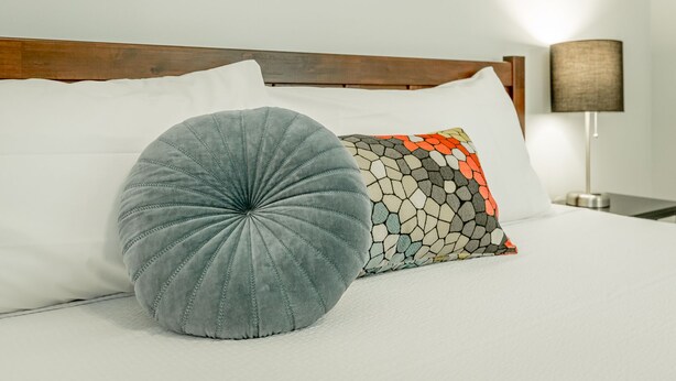 Ein Bett mit weißen Bettwäsche, einem runden grauen Kopfkissen und einem Kissen mit Muster.